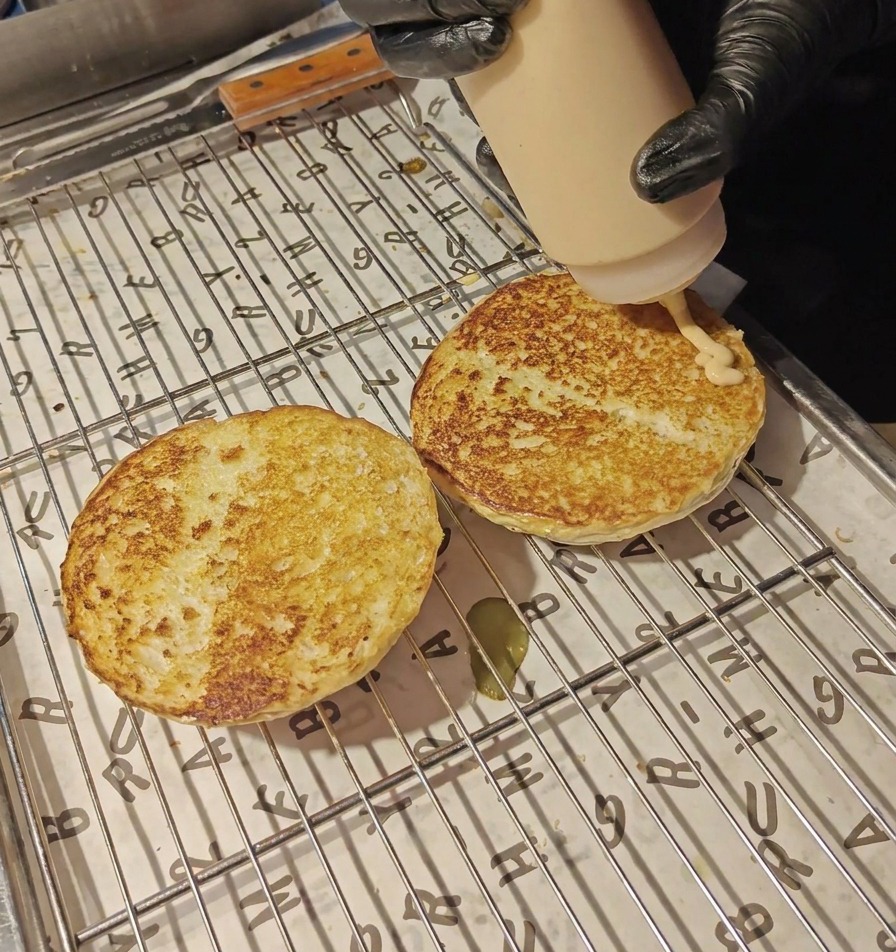 Una salsa cremosa di colore arancione viene versata da un flacone dosatore sulle due metà di un panino tostato posizionate su una griglia.