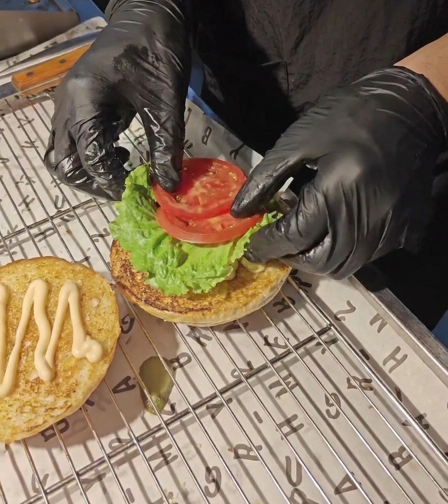 Mani con guanti neri adagiano fette di pomodoro fresco sopra una foglia di lattuga verde sulla base di un panino tostato.