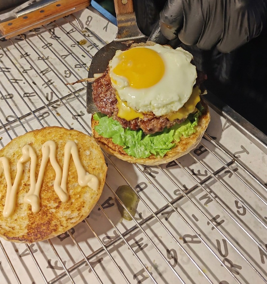 Una spatola metallica trasferisce il patty di manzo con formaggio fuso e uovo allocchio di bue sopra lo strato di lattuga e pomodoro.