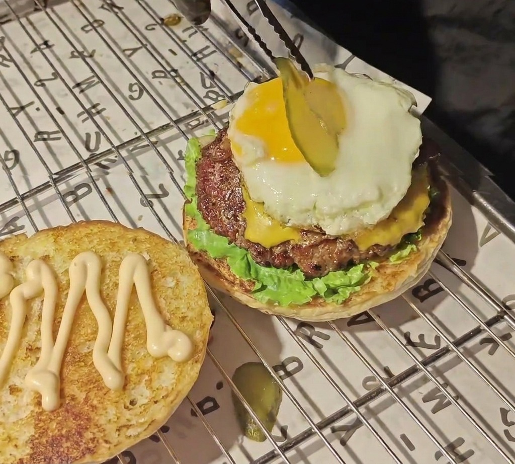 Delle pinze posizionano una sottile fetta di cetriolo sottaceto sopra il tuorlo duovo dorato in cima alla pila del burger.