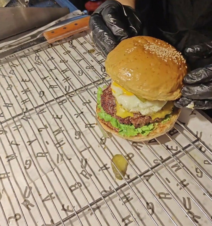 Mani con guanti neri posizionano la parte superiore di un panino dorato ai semi di sesamo su un burger completo con uovo e bacon.