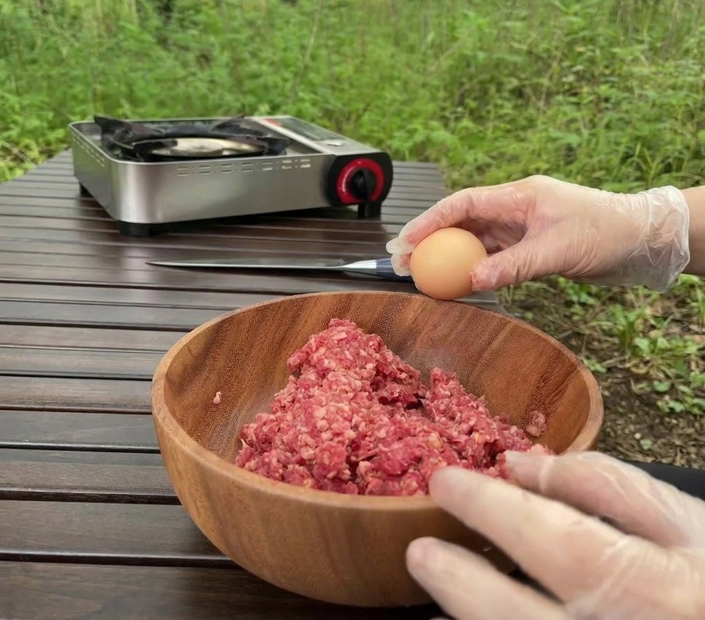 Una persona con guanti trasparenti rompe un uovo in una ciotola di legno piena di carne macinata cruda allaperto.