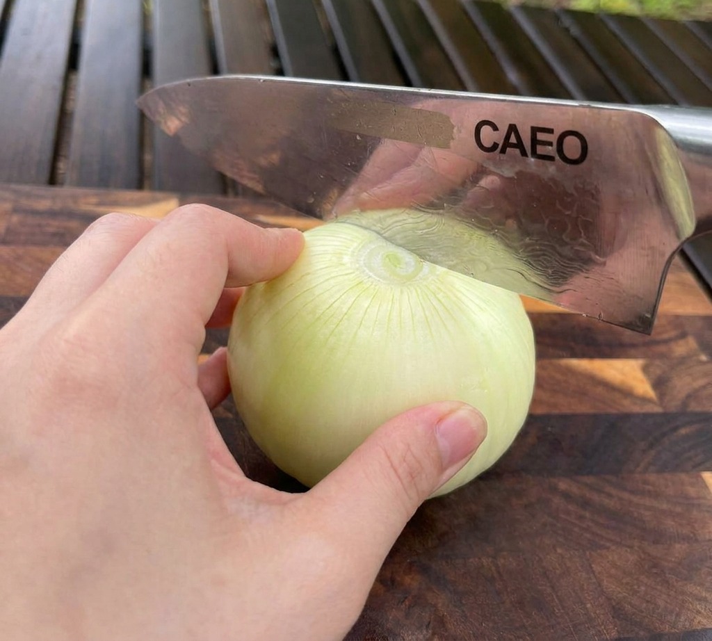 Una mano tiene ferma una cipolla bianca su un tagliere mentre un coltello da chef la affetta.