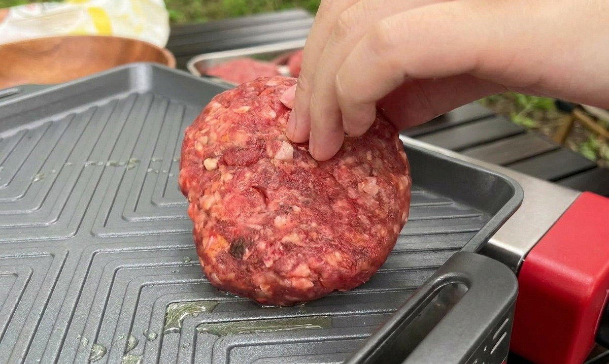 Una mano posiziona uno spesso hamburger di carne cruda su una piastra quadrata in ghisa molto calda.