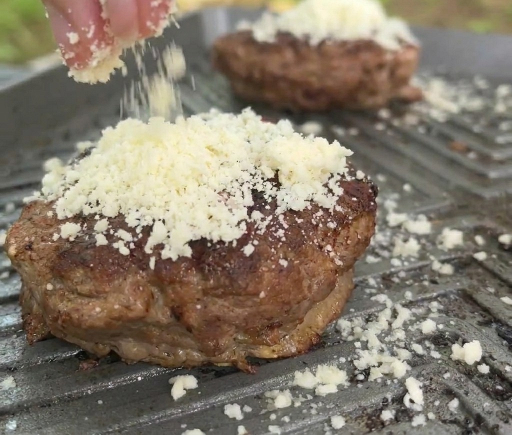 Dettaglio di formaggio bianco grattugiato cosparso su un hamburger di manzo in cottura sulla piastra.