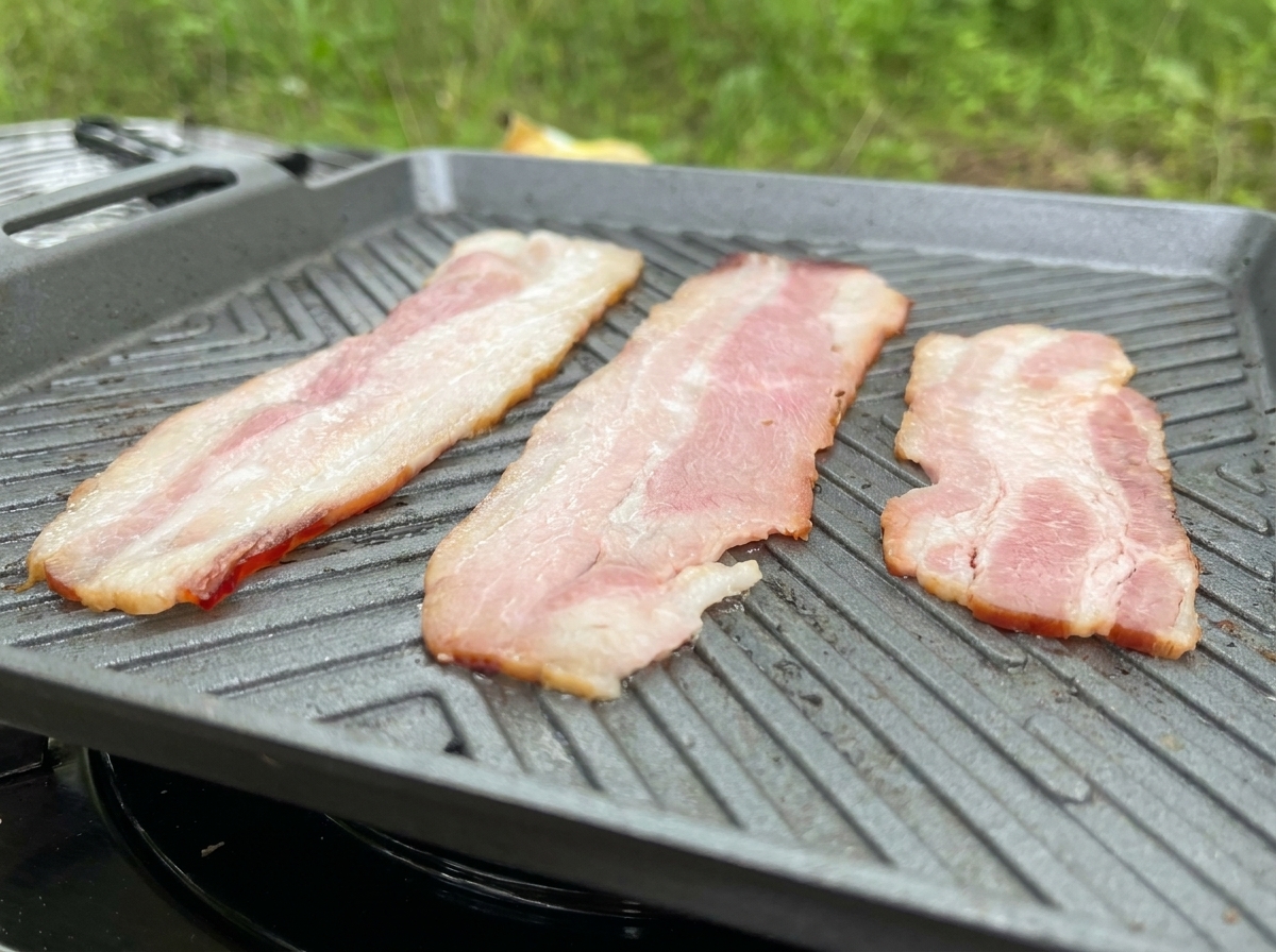Tre lunghe fette di bacon sfrigolano su una piastra grigia, rilasciando il loro grasso.