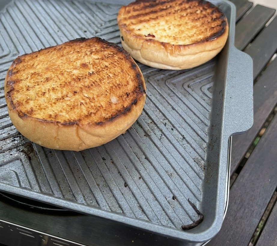 Un cucchiaio dargento spalma una densa salsa rosa sulla superficie dorata di un panino brioche tostato.