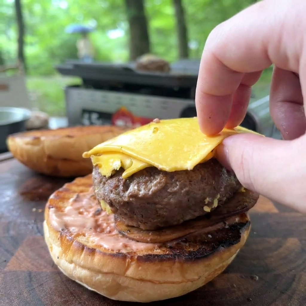 Una mano posiziona un hamburger cotto con formaggio giallo fuso su un panino tostato e spalmato di salsa rosa.