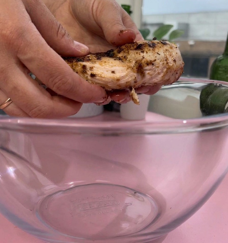 Mani che adagiano un pezzo di petto di pollo cotto e condito in una ciotola capiente di vetro trasparente su una superficie rosa.