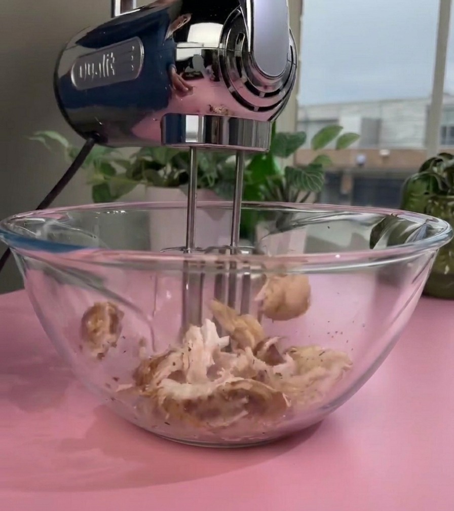 Uno sbattitore elettrico con le fruste in movimento che sfilaccia il pollo cotto in una ciotola di vetro creando una consistenza finissima.
