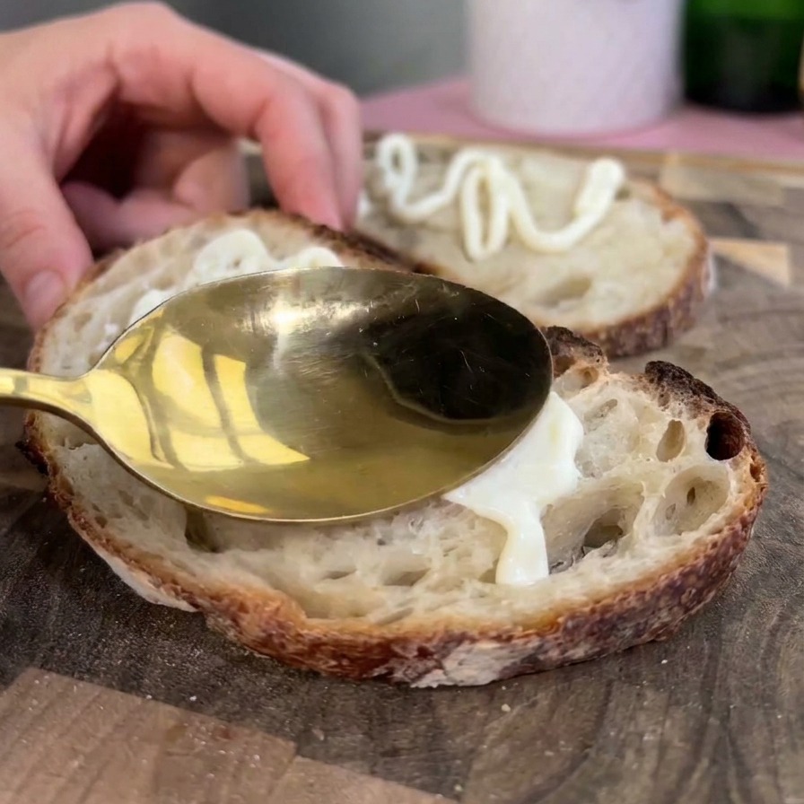 Un cucchiaio dorato che spalma un generoso strato di maionese bianca su una fetta tostata di pane rustico sourdough.