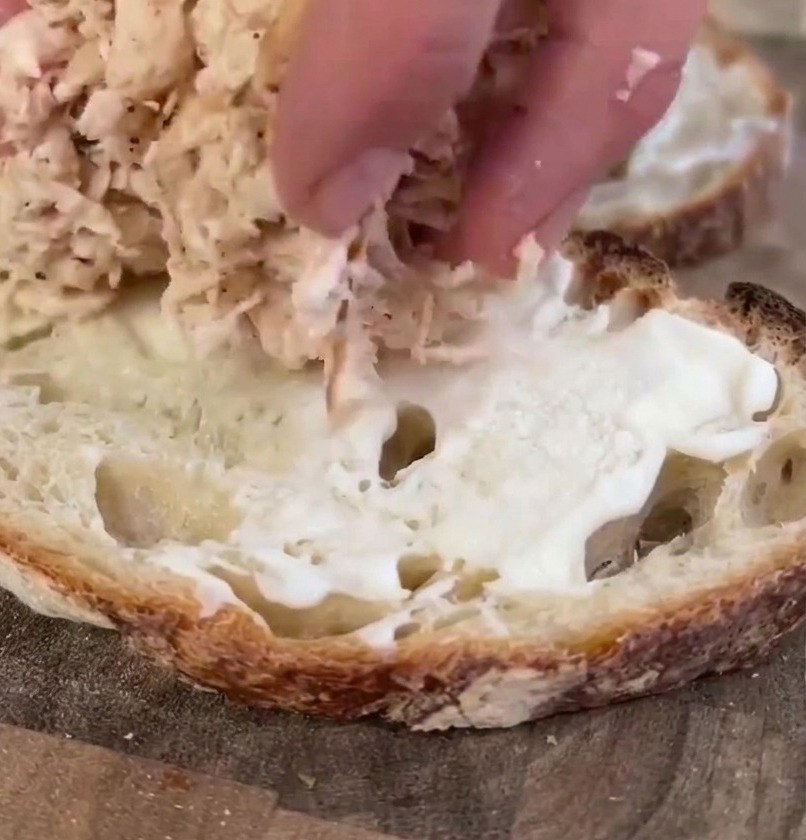 La mano di una persona che adagia una generosa porzione di pollo cotto sfilacciato su una fetta di pane sourdough già spalmata di maionese.