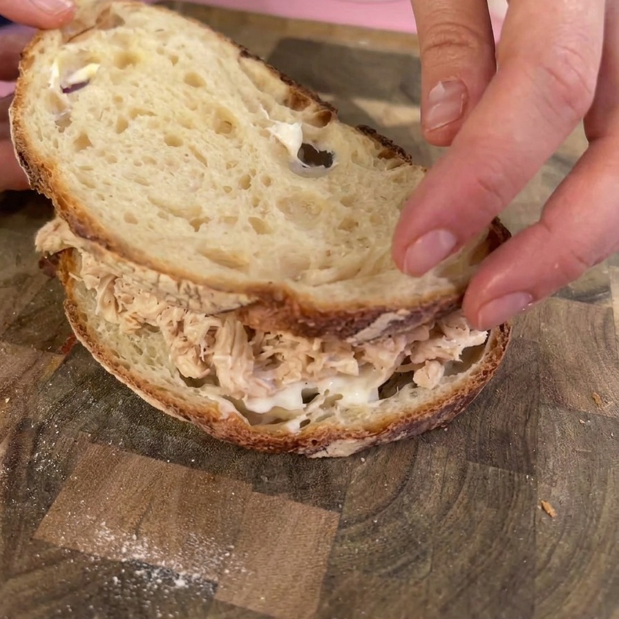 Mani che posizionano una fetta tostata di pane sourdough sopra una ricca porzione di pollo sfilacciato per chiudere il panino.