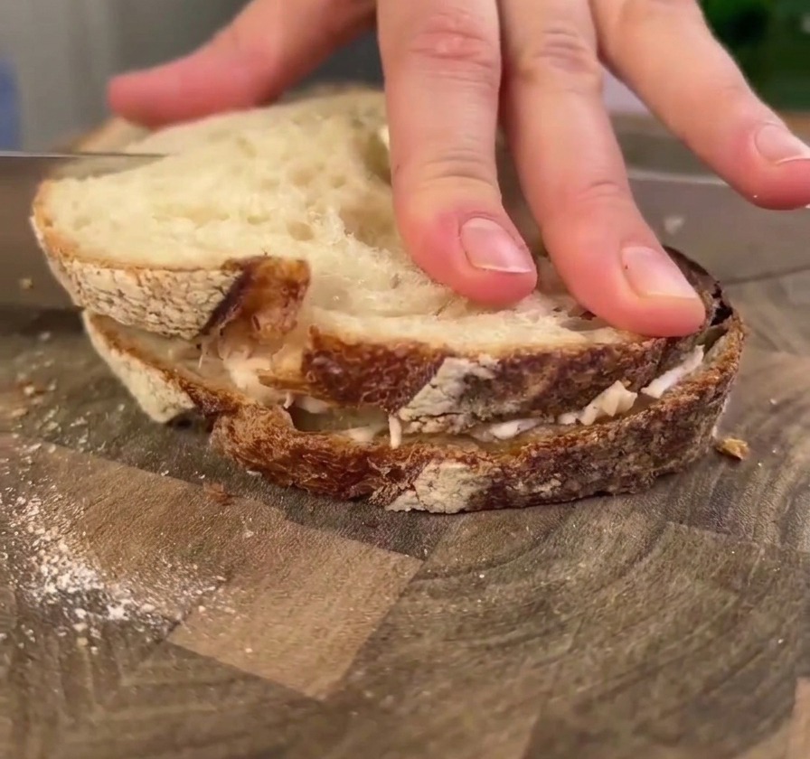 Una mano che esercita una pressione decisa su un grande panino al pollo e pane sourdough su un tagliere di legno, mentre un coltello si appresta a tagliarlo.