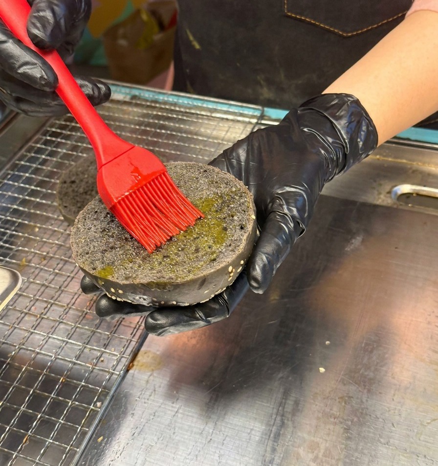 Una mano con guanto nero usa un pennello in silicone rosso per spennellare dolio linterno di un panino nero al carbone con sesamo.