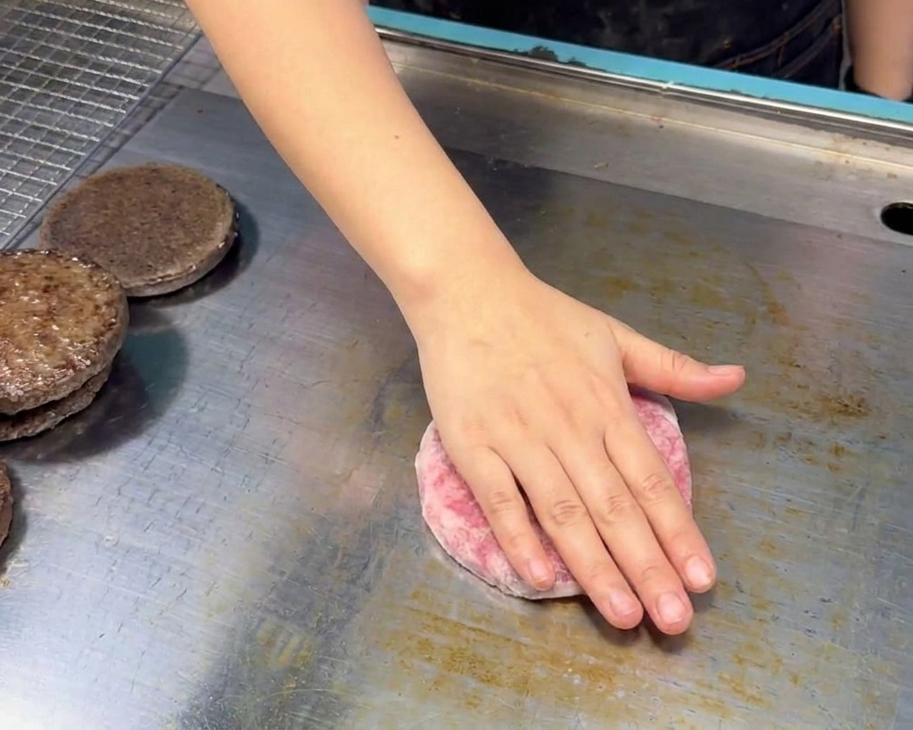 Una mano adagia un tortino di carne di manzo cruda e rosa su una piastra in acciaio inossidabile pulita accanto alle metà del panino tostato.