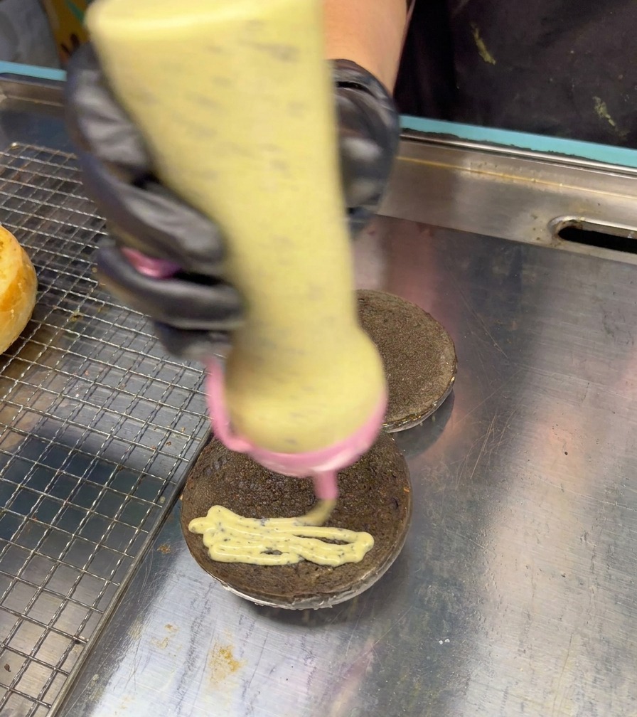 Un flacone dosatore distribuisce una salsa cremosa e punteggiata al tartufo con un movimento a zig-zag su un panino nero.