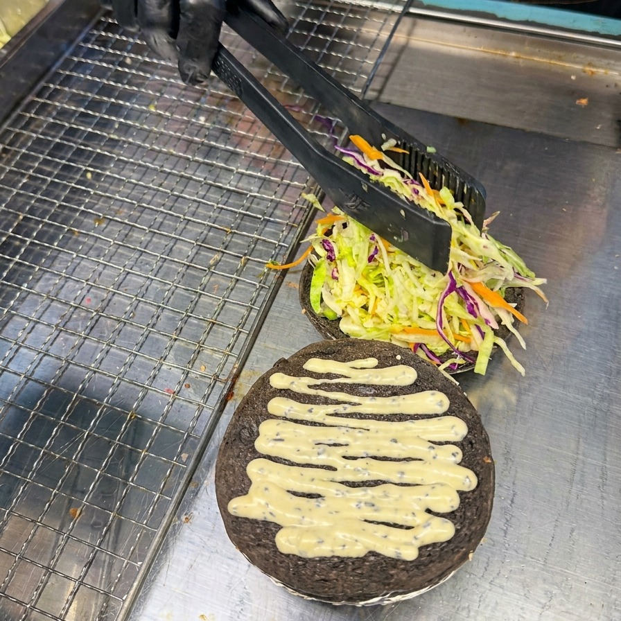 Una persona usa le pinze per posizionare con cura una fetta di pomodoro rosso vivo sul letto di verdure in un panino al carbone.