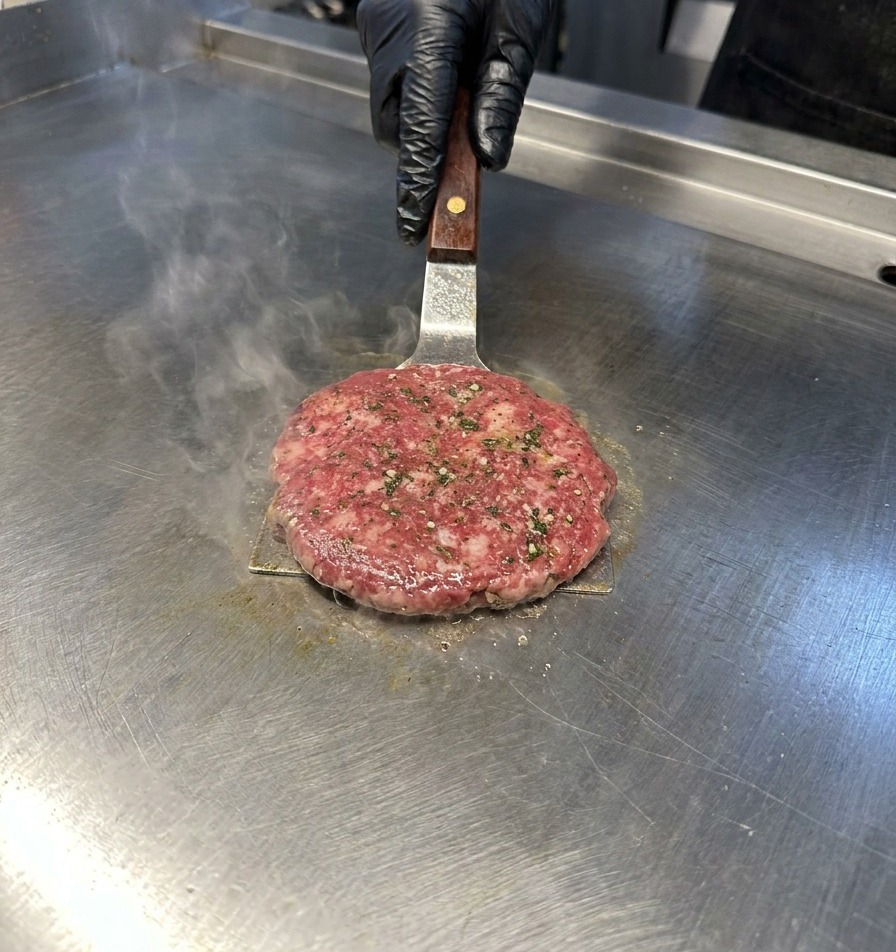 Una spatola di metallo preme su un tortino di carne su una piastra in acciaio inossidabile da cui sale del vapore.