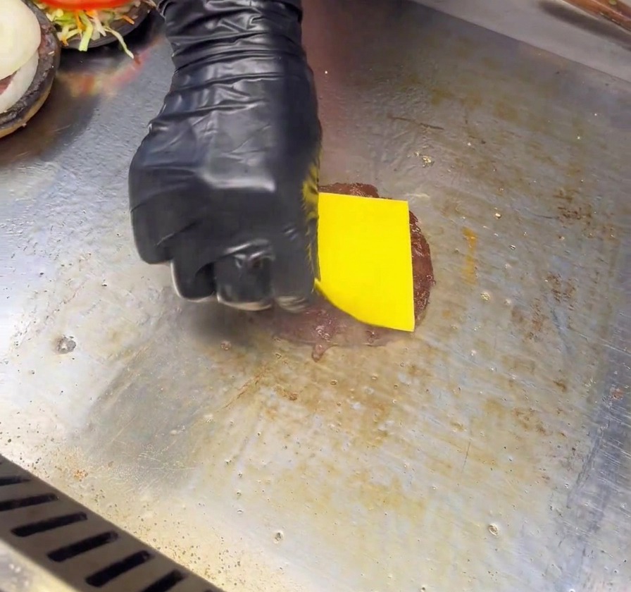 Una mano con guanto nero posiziona una fetta di formaggio giallo su un hamburger scottato su una piastra liscia.