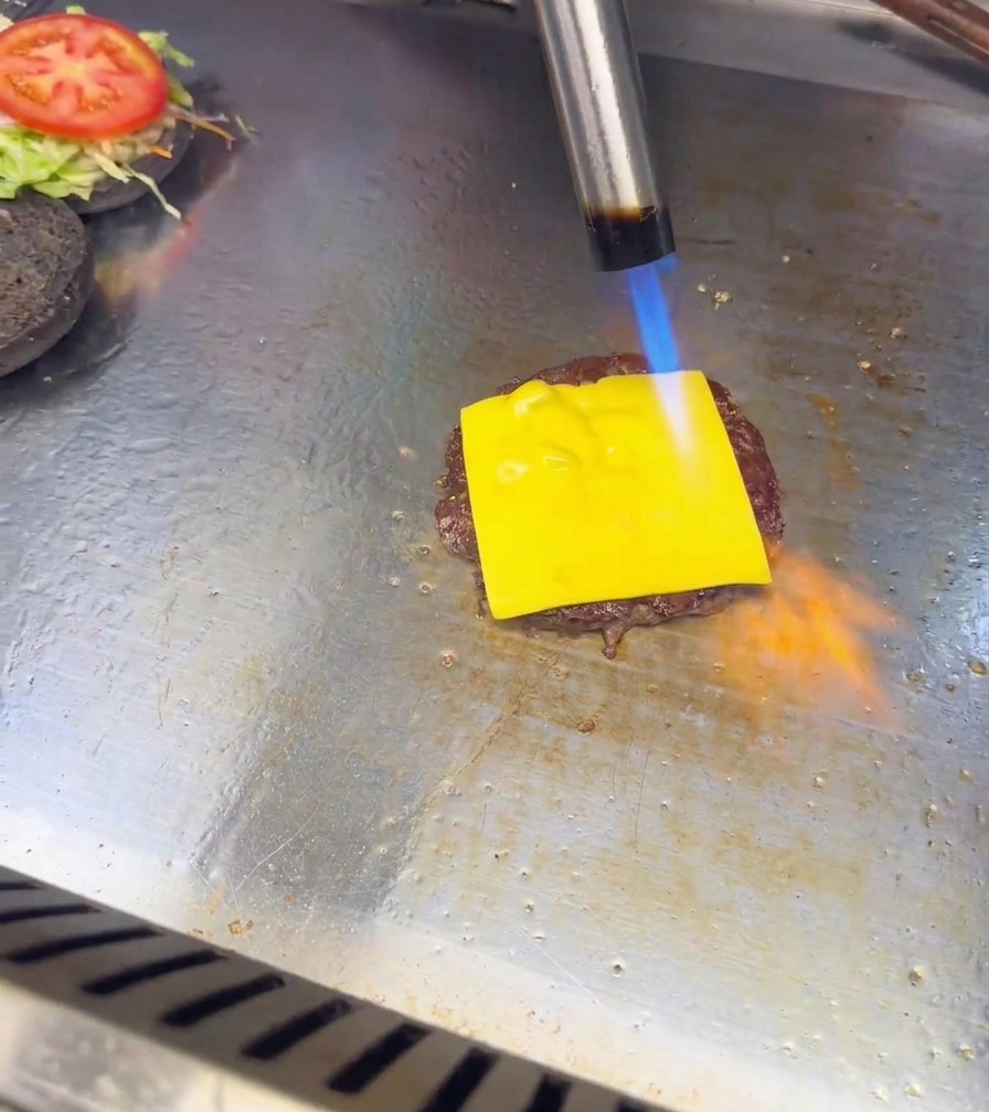 La fiamma blu e arancione di un cannello da cucina fonde una fetta di formaggio sopra un tortino di carne per burger.
