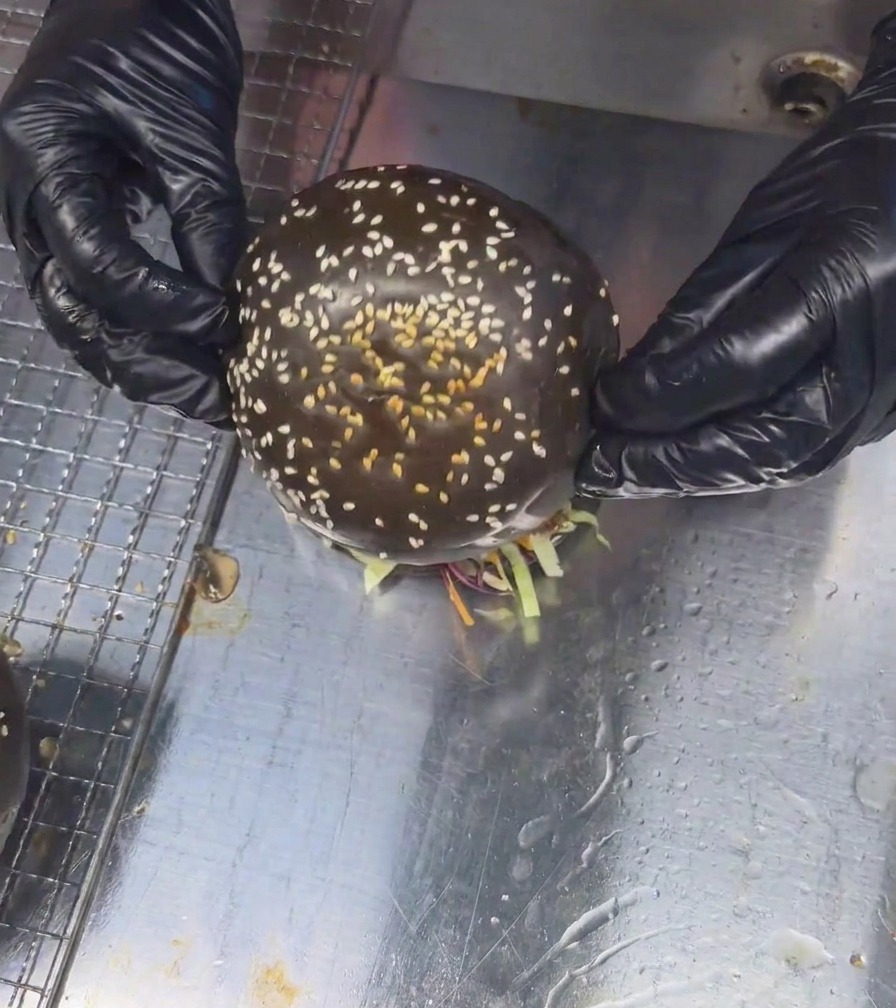 Due mani con guanti posizionano la calotta superiore di un panino nero con sesamo su un imponente burger ai funghi finito.
