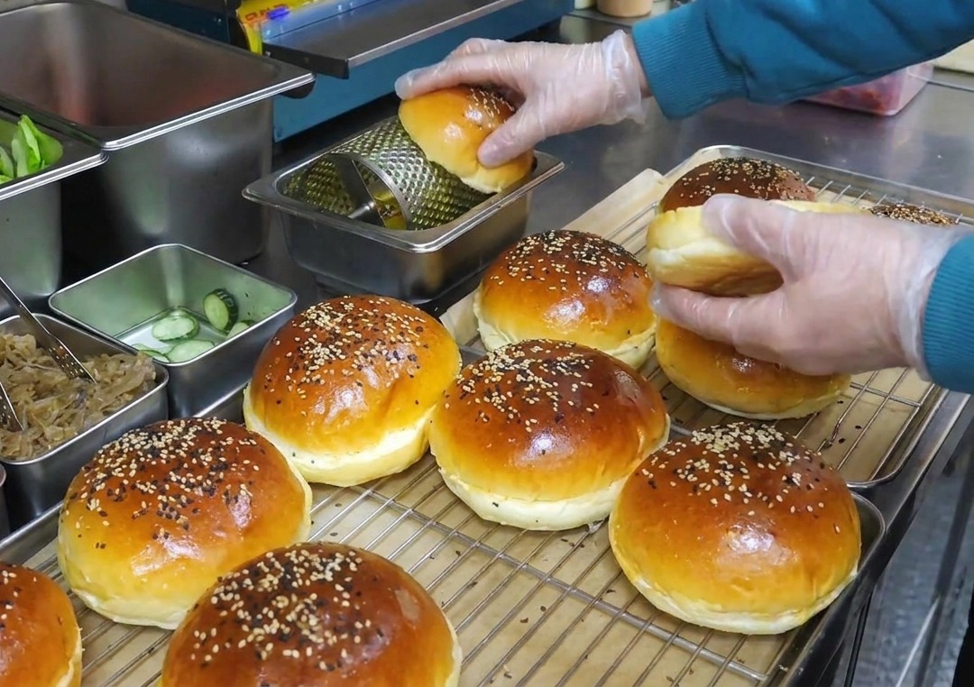 Mani con guanti di plastica che fanno girare un panino da burger su un rullo dosatore di burro in metallo.