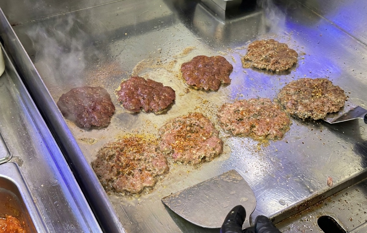 Patty di carne per burger che vengono schiacciati e scottati su una piastra in acciaio con una spatola metallica.