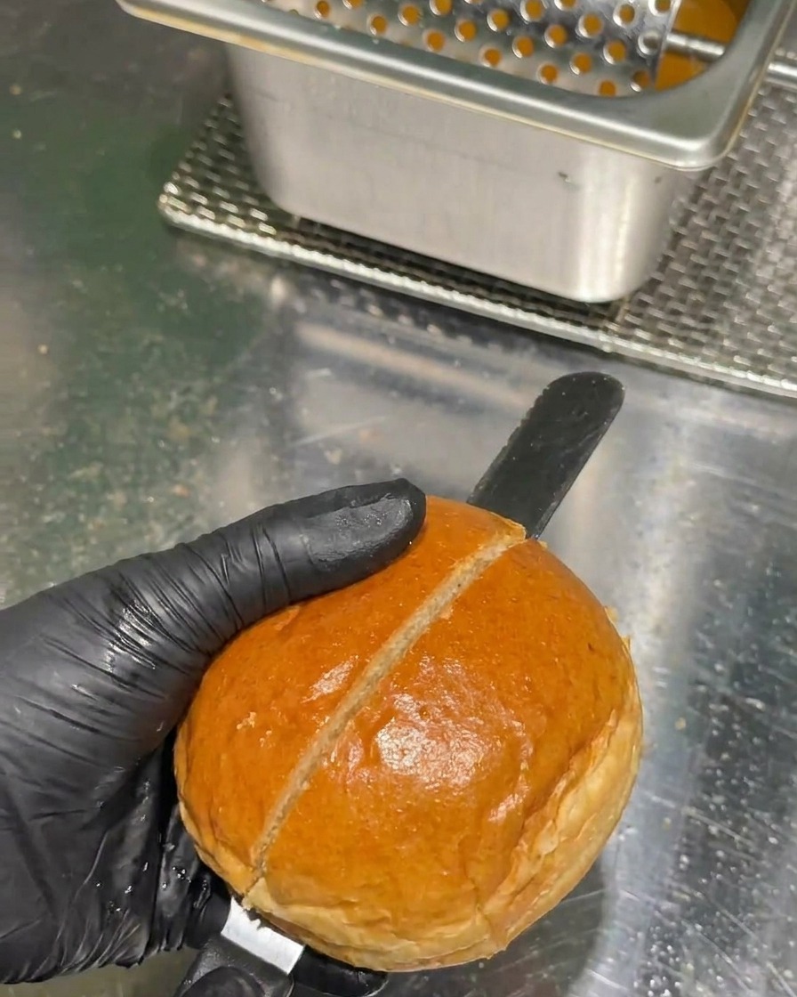 Una mano con un guanto nero usa un lungo coltello seghettato per tagliare a metà un panino dorato da burger su un bancone in acciaio inossidabile.