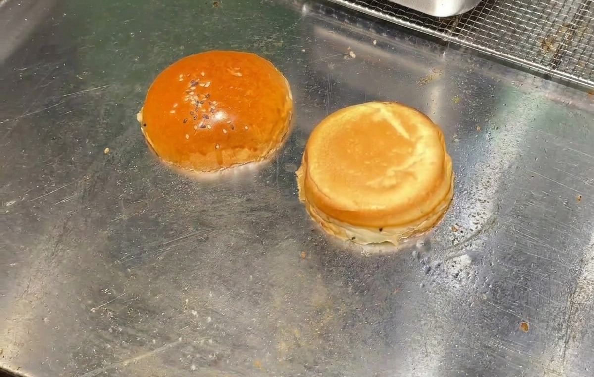 Le due metà di un panino da burger che tostano rivolte verso il basso sulla superficie di una piastra metallica liscia.