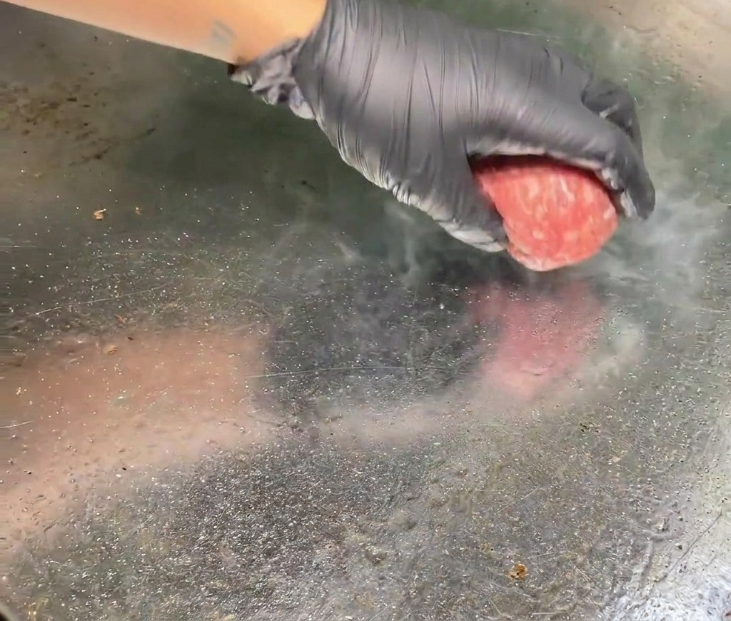 Una mano con un guanto posiziona una polpetta di manzo cruda e rosa su una piastra metallica bollente con vapore visibile che sale dal punto di contatto.