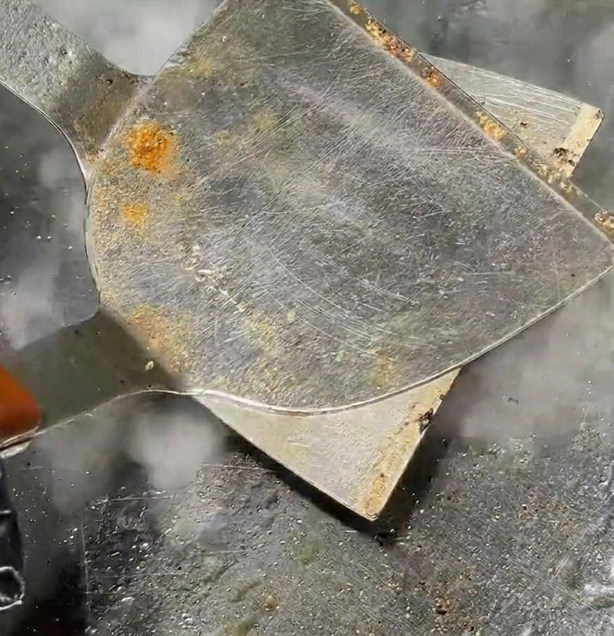 Una spatola metallica che schiaccia una polpetta rotonda trasformandola in un tortino sottile su una piastra calda con vapore che sale.