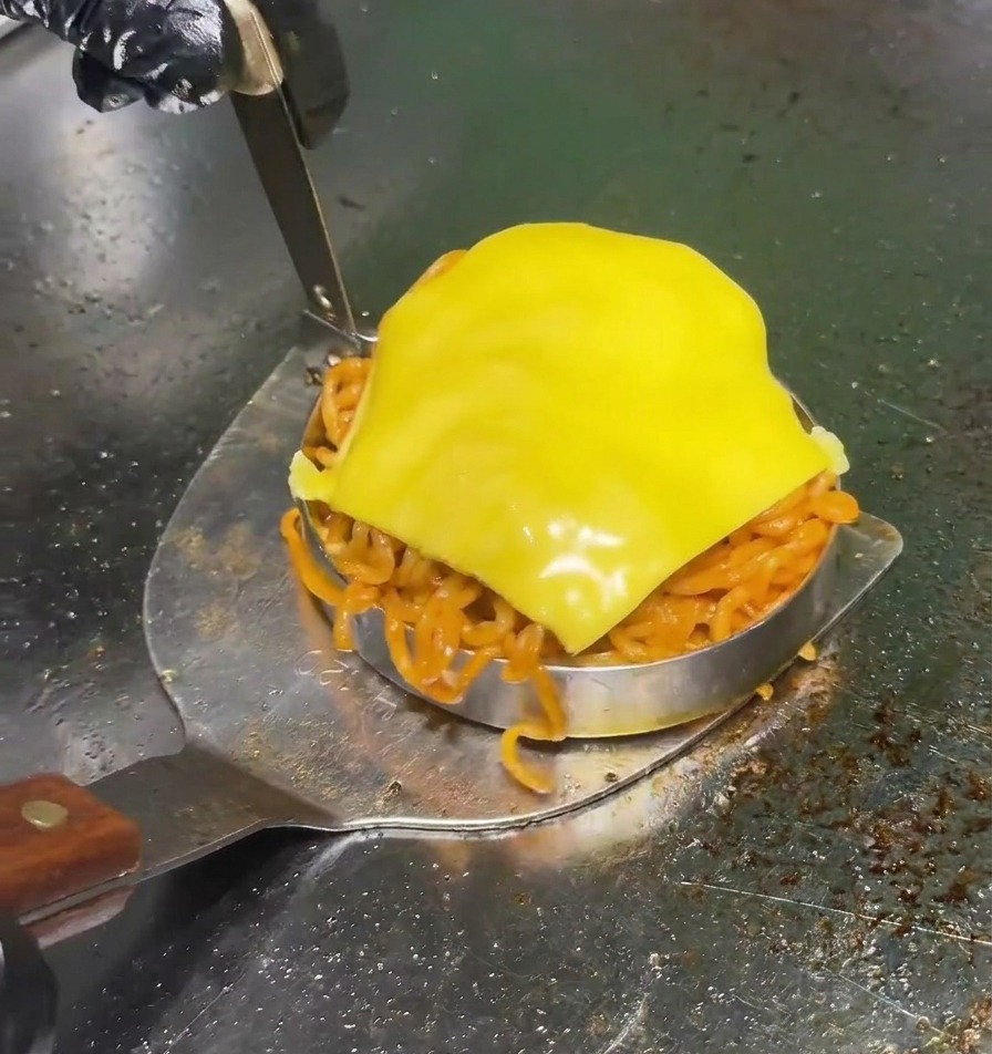Una spatola metallica che solleva un coppapasta circolare riempito di noodle rossi piccanti e sormontato da una fetta di formaggio giallo su una piastra professionale.