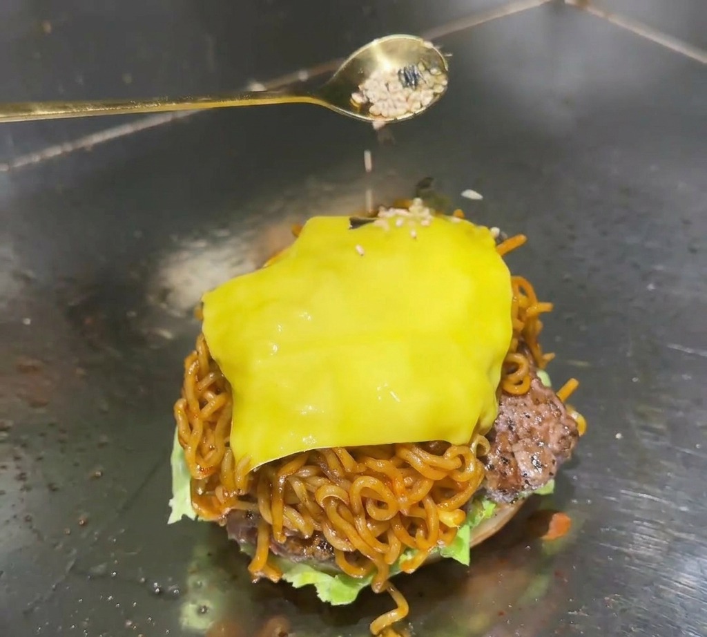 Un cucchiaio dorato che distribuisce una miscela di semi di sesamo bianco e fiocchi di alga nera sul formaggio fuso in cima a un ramen burger.