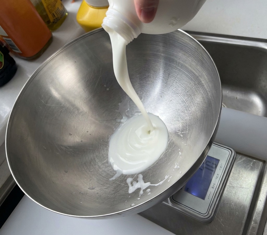 Yogurt bianco versato da una confezione di plastica in una grande ciotola in acciaio inossidabile.
