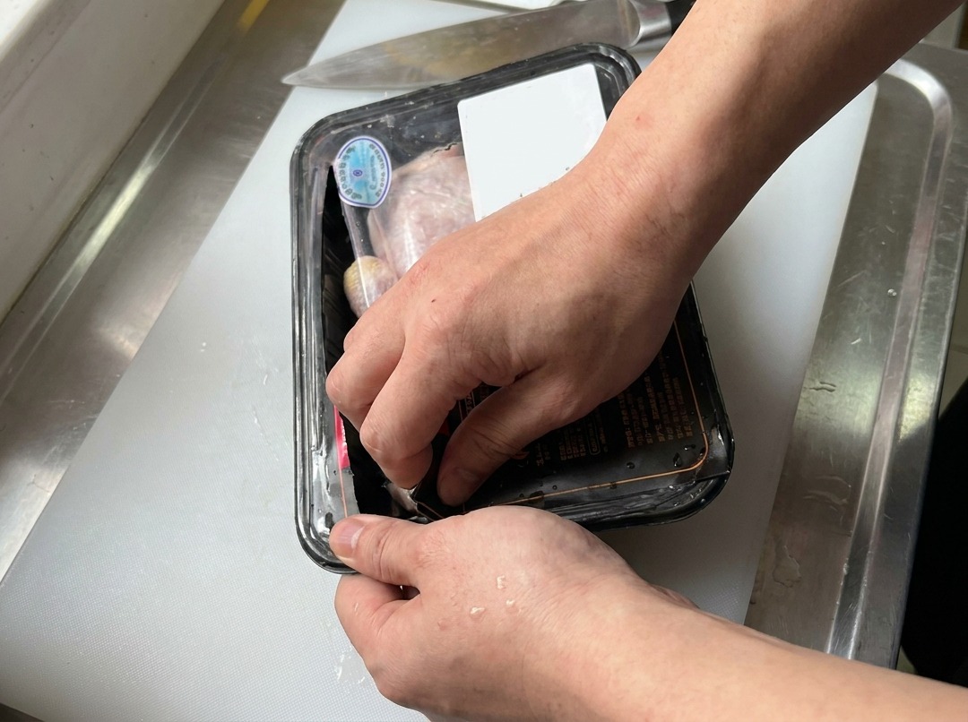 Mani che aprono una vaschetta di sovracosce di pollo crude su un tagliere bianco accanto a un coltello da cucina.