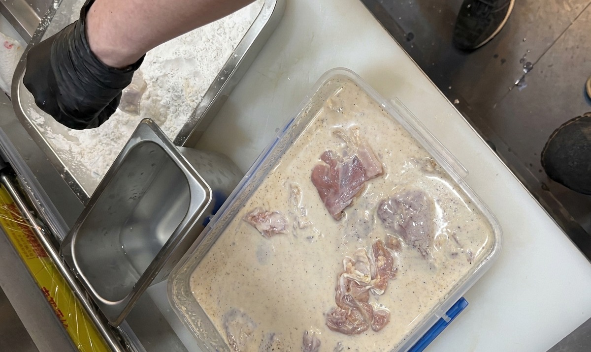 Mani con guanti che trasferiscono il pollo marinato dal liquido cremoso a un vassoio di amido e farina speziati.
