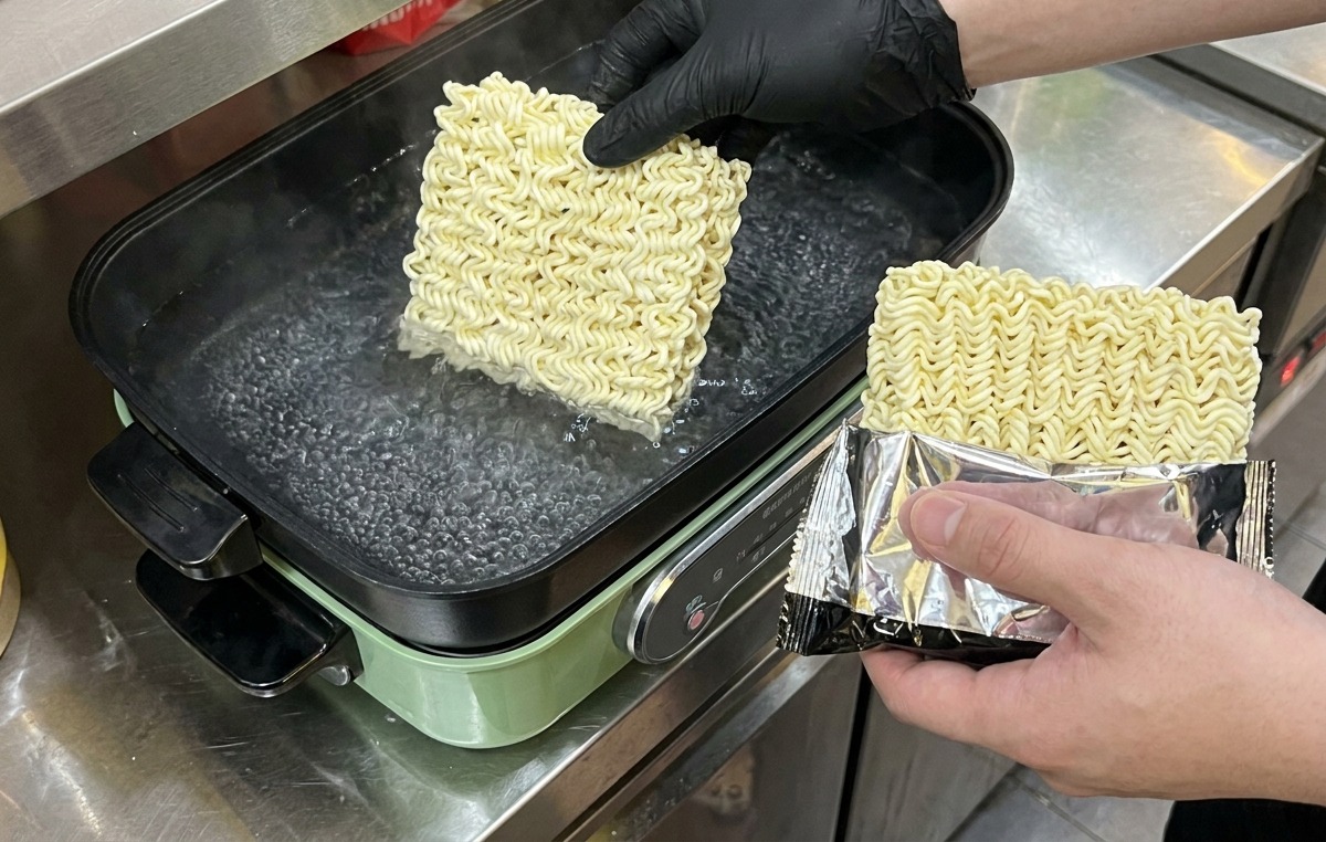 Una persona con guanti neri che immerge i blocchi quadrati di noodle istantanei in una pentola di acqua bollente.
