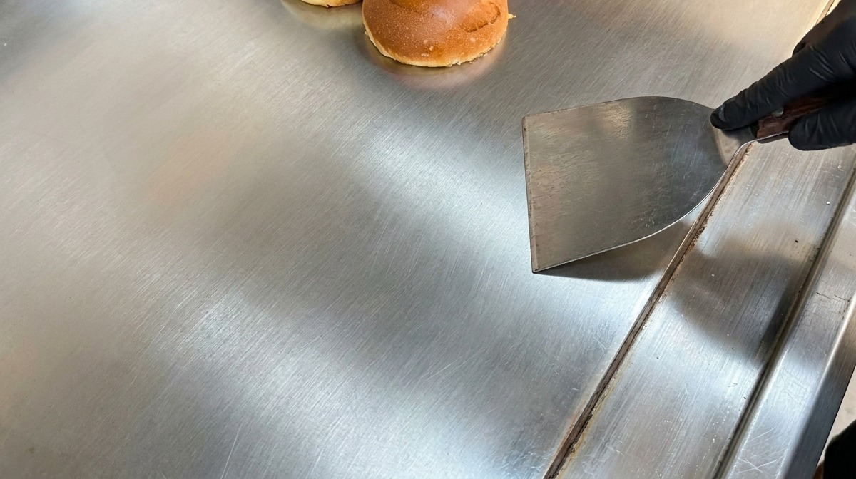 Panini brioche tagliati a metà che tostano su una grande piastra in acciaio accanto a una spatola di metallo.