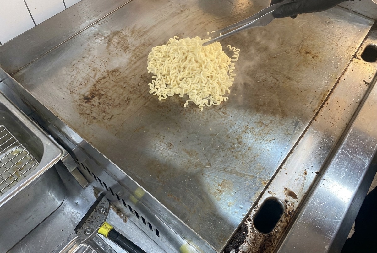 Noodle fumanti e burro che si scioglie su una grande superficie di cottura in metallo lucido.