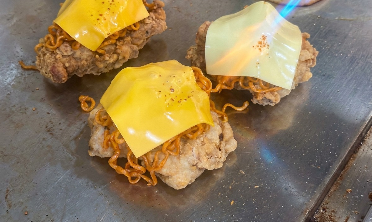 La fiamma di un cannello da cucina che scioglie una fetta di formaggio giallo sopra una pila di noodle piccanti e pollo fritto.