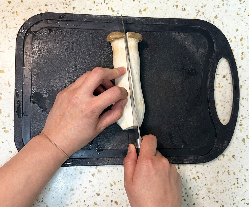 Una persona che usa un coltello da chef per tagliare a metà per il lungo un grande fungo cardoncello su un tagliere di plastica nera.
