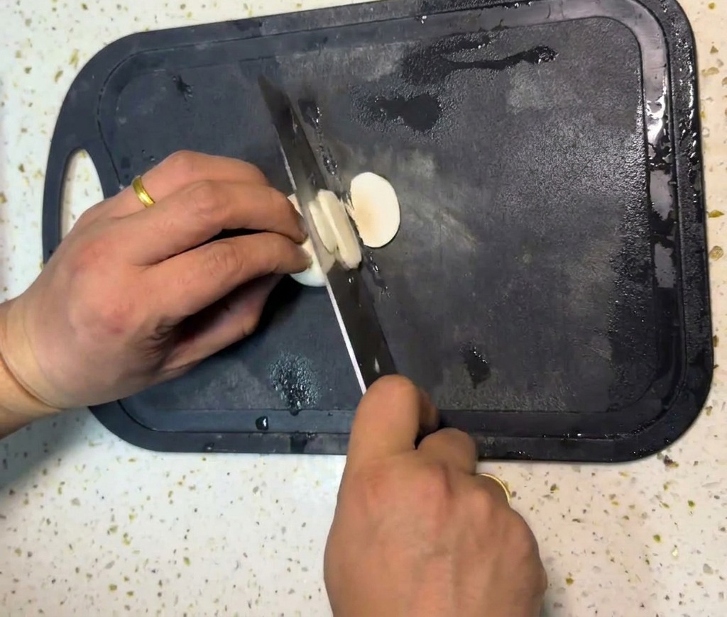 Le mani di una persona che usano un coltello da chef per tagliare i funghi champignon bianchi a fette sottili su un tagliere nero.