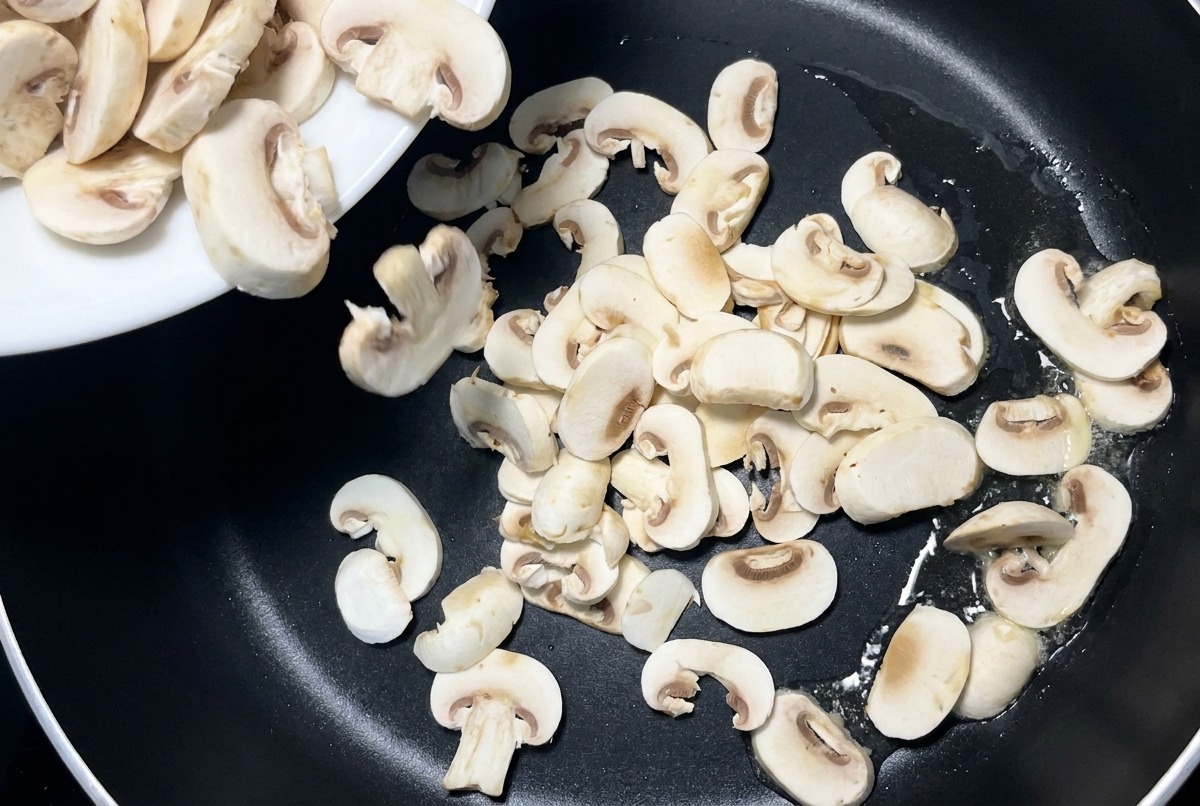 Funghi bianchi affettati che vengono versati da un piatto di ceramica bianca in una padella antiaderente nera.