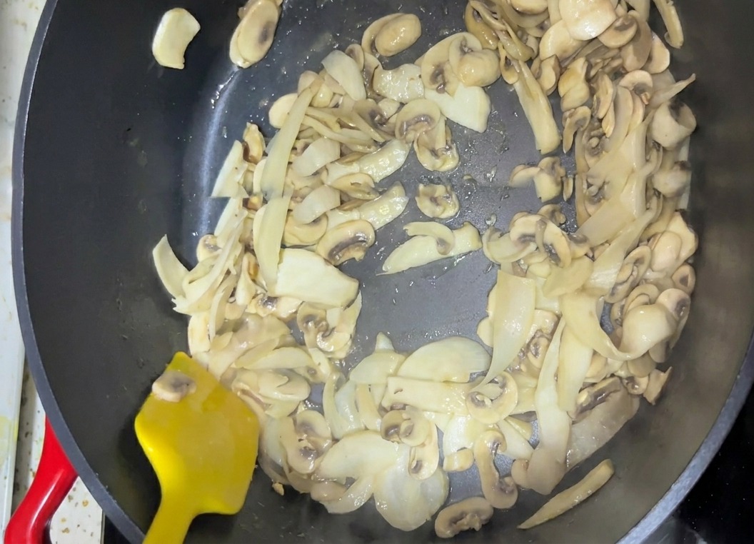 Funghi affettati che vengono mescolati in una padella scura con una spatola giallo brillante.