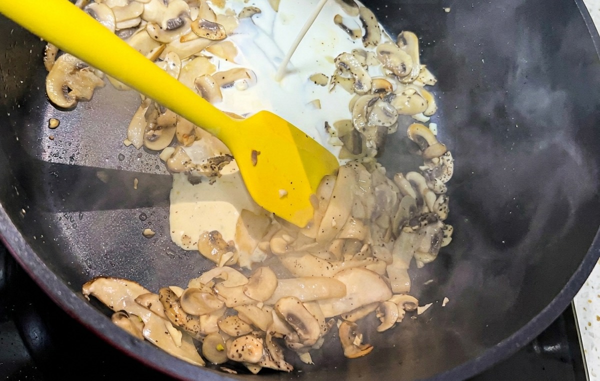 Un filo di panna fresca liquida che viene versato in una padella nera piena di funghi saltati a fette.