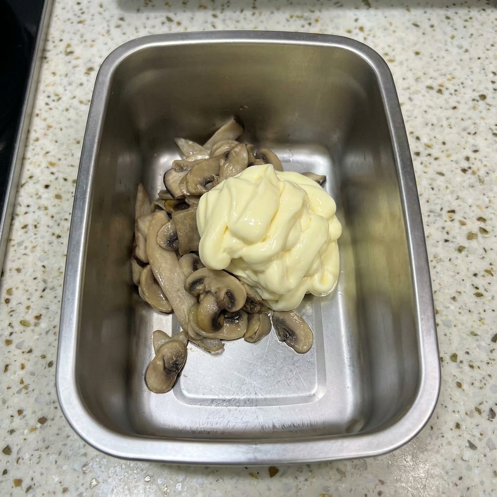 Un contenitore di metallo con funghi saltati sormontati da un grande ciuffo di maionese giallo pallido.