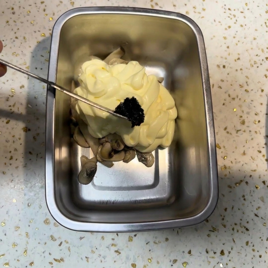 Un piccolo utensile di metallo che deposita una porzione di salsa al tartufo nero scuro su una montagnola di maionese e funghi.