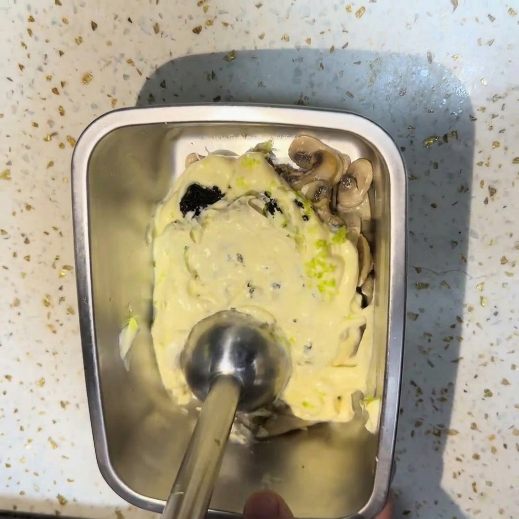 Un frullatore a immersione che amalgama funghi saltati, panna e salsa al tartufo nero in un contenitore di metallo per creare una salsa liscia e densa.