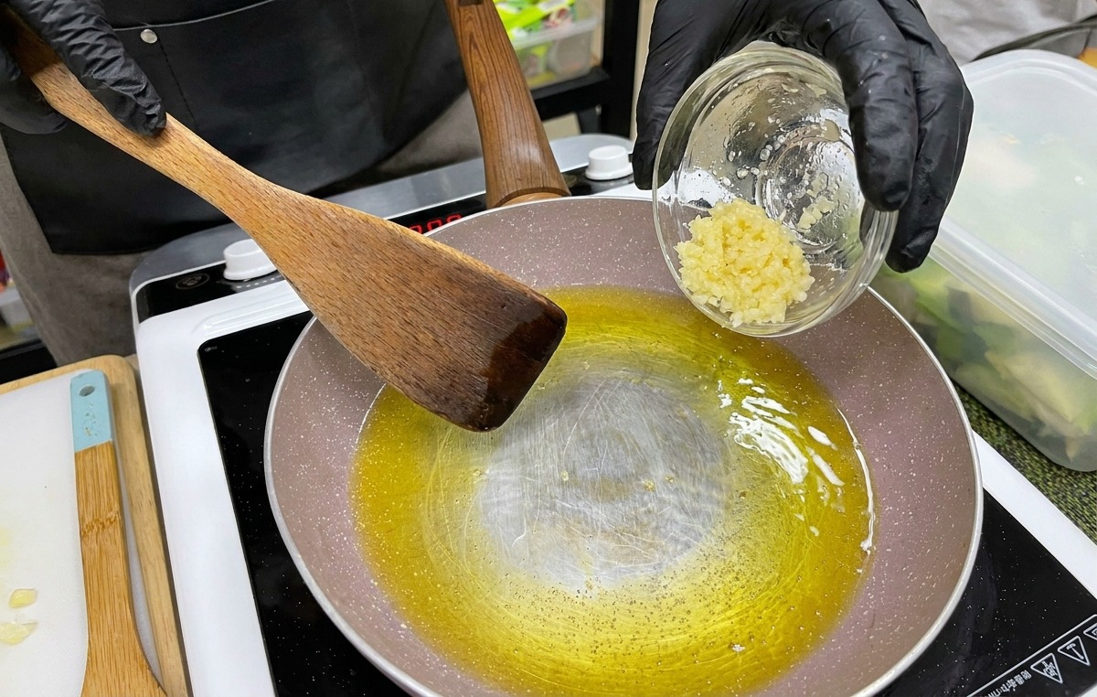 Una mano con un guanto nero versa una ciotolina di aglio tritato in una padella con burro fuso e olio.